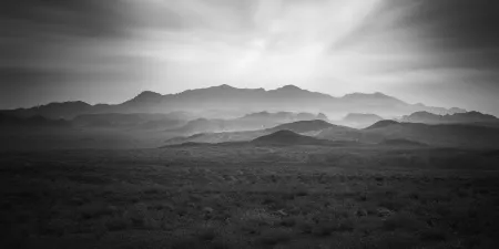 A series of dark, overlapping mountain ranges under a bright, cloudy sky in black and white.
